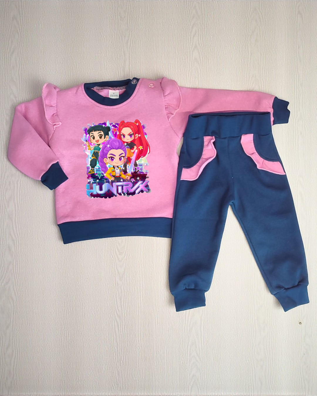 Conjunto de roupa de criança rosa e azul com estampado de personagens e texto HUNTR/X