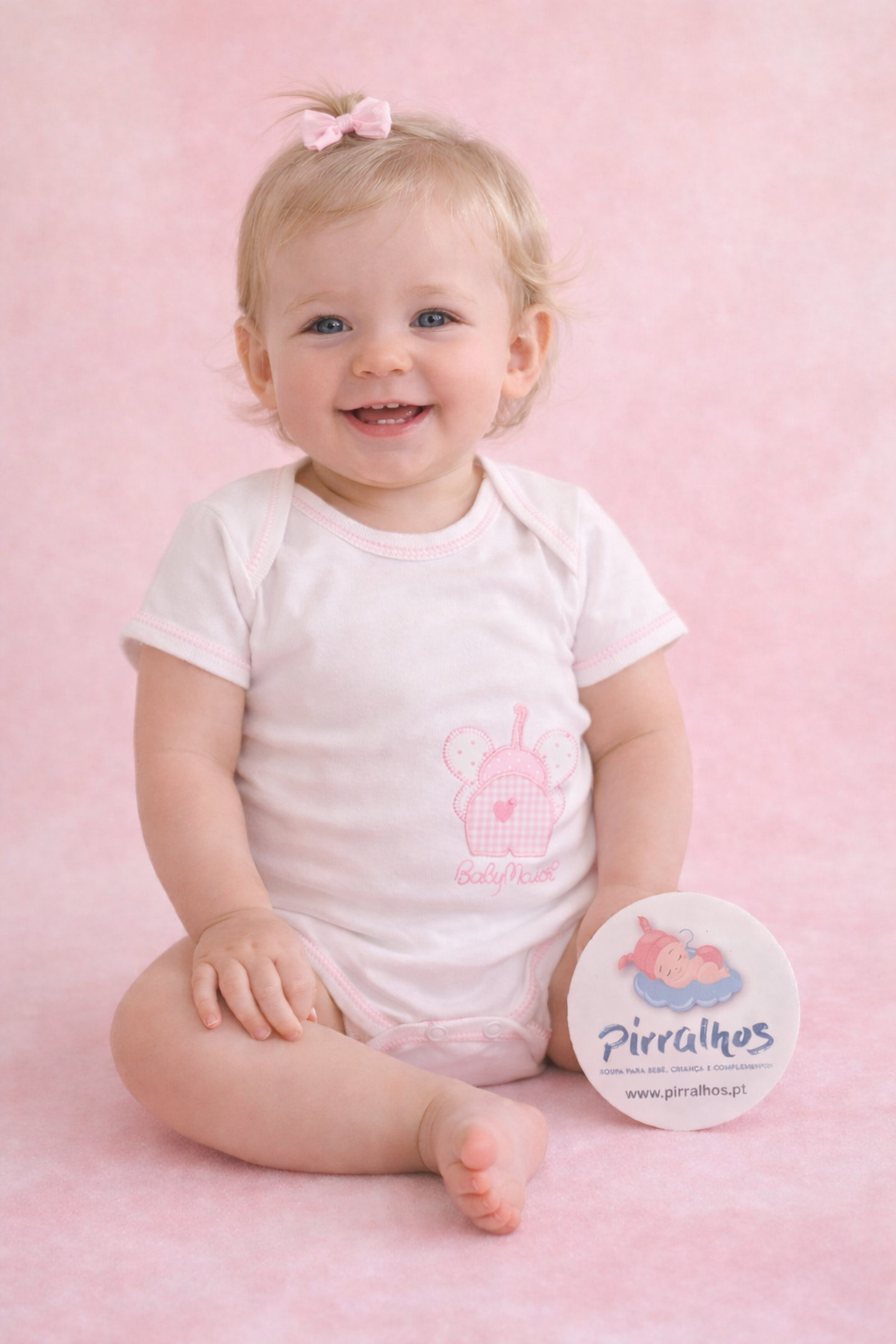 Bebé com body branco e rosa segurando círculo com logo Pirralhos sentado em fundo rosa