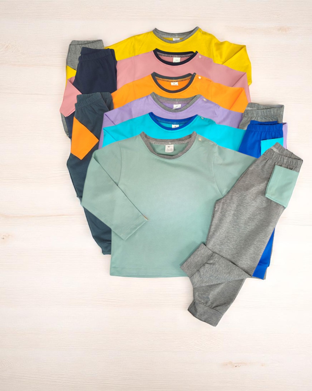 Conjunto de pijamas infantis coloridos, camisolas manga comprida e calças, com bolso em material cinza e verde, em fundo claro.