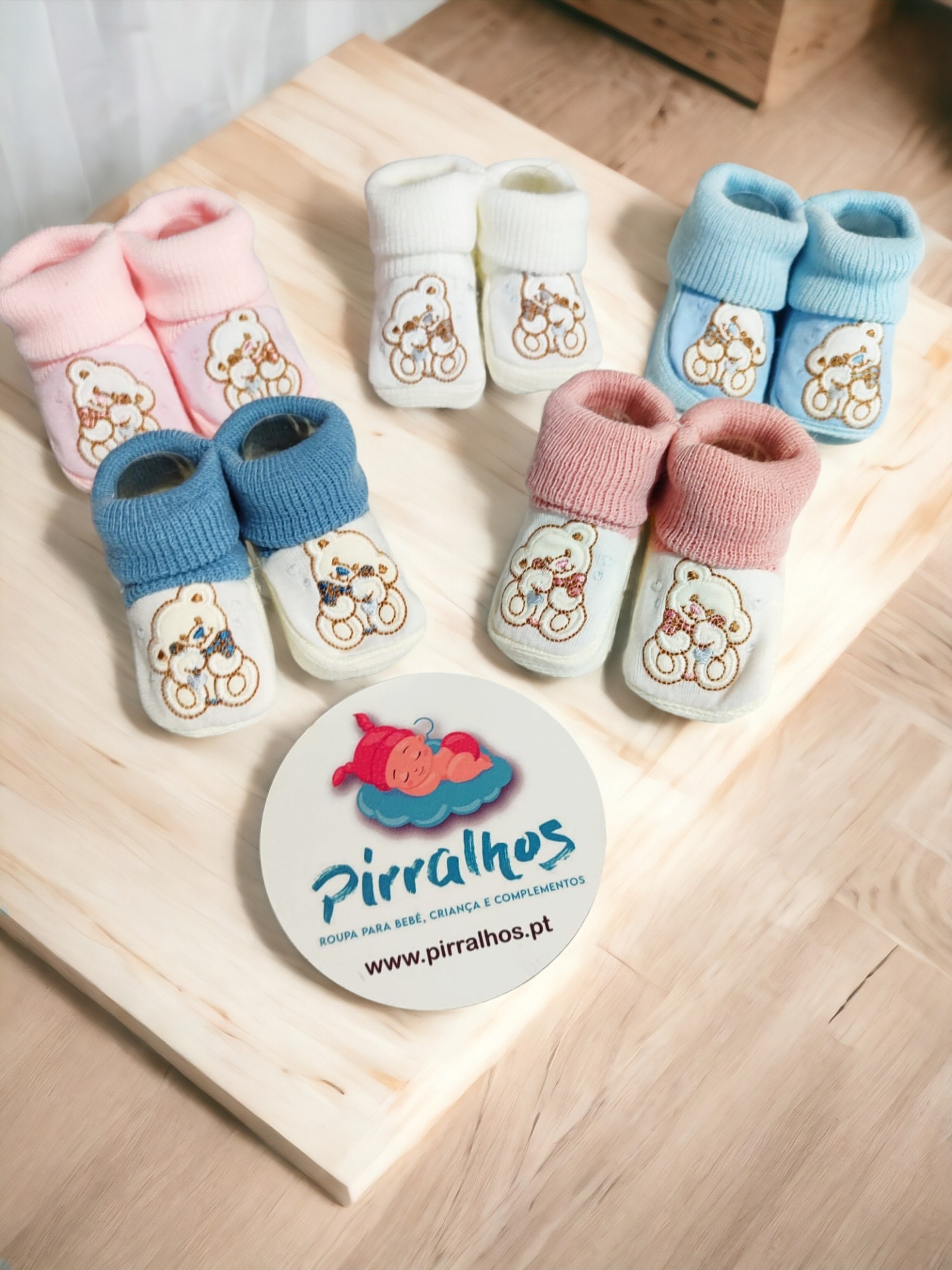 Cinco pares de botins para bebé em várias cores com ursinhos bordados, sobre cadeira de madeira clara e logotipo da marca Pirralhos.
