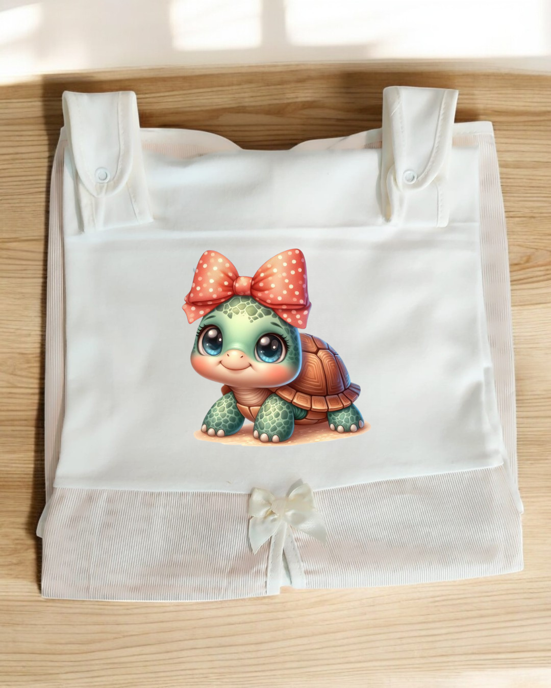 Saco branco com estampa de tartaruga verde de olhos grandes e laço coral, sobre superfície de madeira clara