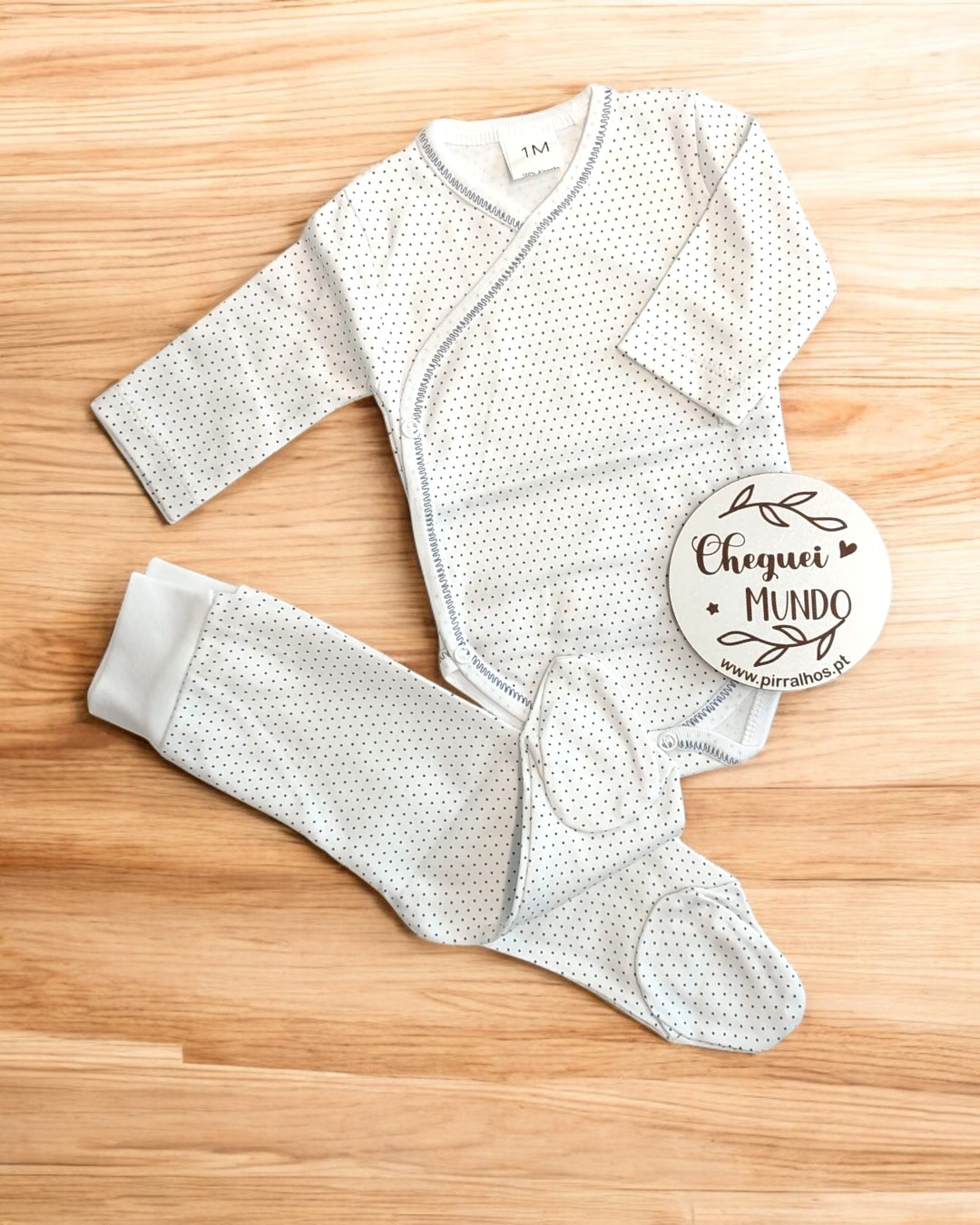roupa de bebé branca com pontos pretos e placa CHEGUEI MUNDO sobre chão de madeira