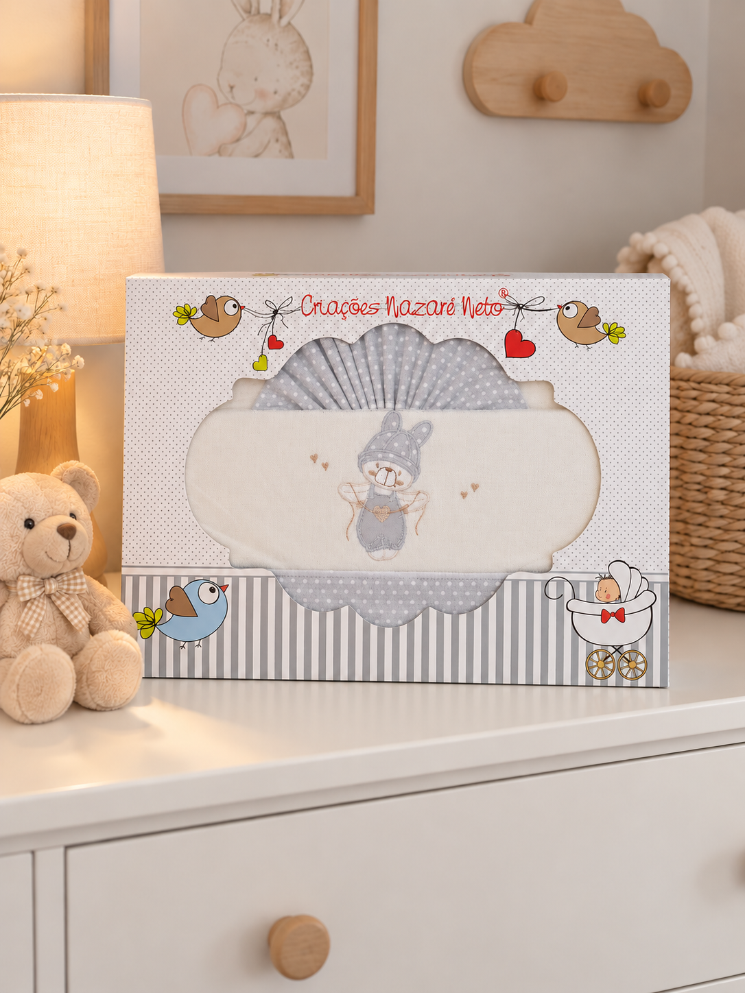 Conjunto de têxteis para bebé cinza e bege numa caixa decorada sobre cómoda branca com peluche e abajur