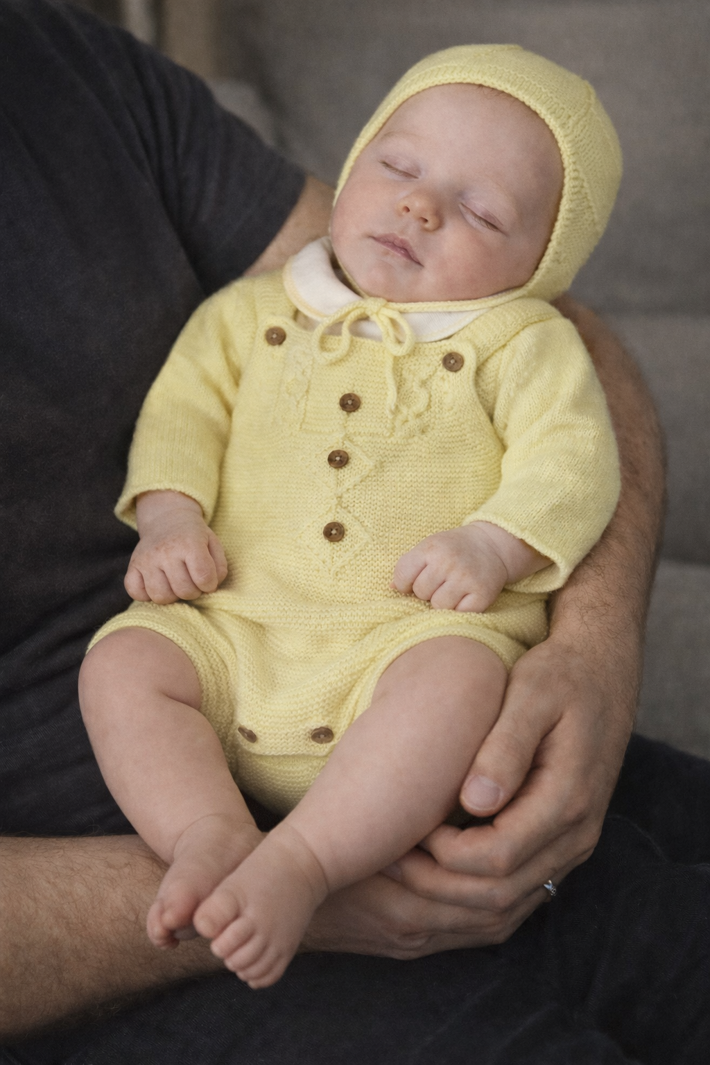 Bebé com macacão e capuz amarelos dormindo no colo de adulto com camiseta cinza escuro