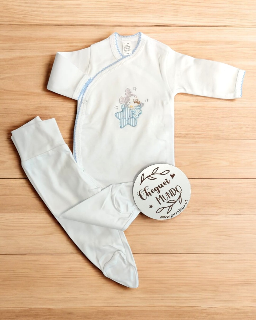 Pijama de bebé branco com ursinho numa estrela azul, fecho lateral e detalhes azuis, sobre madeira clara, com plaqueta redonda branca.