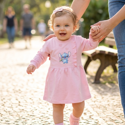 Bebé com vestido rosa e sapatinhos a caminhar ao ar livre com ajuda de um adulto