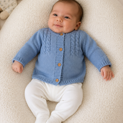 Bebé vestindo casaco azul de malha com botões de madeira, deitado em almofada creme com urso de peluche ao lado.