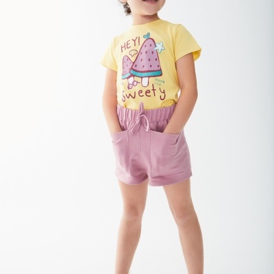 Criança com t-shirt amarela, calções rosa e sapatos brancos em fundo branco