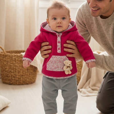 Bebé com camisola rosa, calças cinza e ursinho de peluche, com adulto ao lado em ambiente interior claro