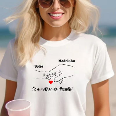 Pessoa com t-shirt branca estampada com mãos desenhadas e texto em português, óculos de sol e copo azul claro na mão, cenário de praia