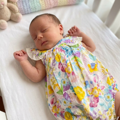 Bebé a dormir vestido com macacão floral colorido ao lado de coelho de peluche e manta de tricô colorida.