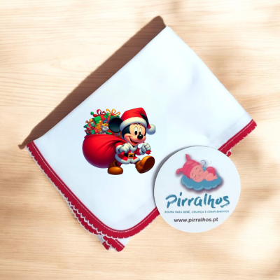 Fralda branca com bordas vermelhas e estampa de Mickey Mouse de Pai Natal com saco de presentes, sobre superfície de madeira clara com etiqueta redonda ao lado.