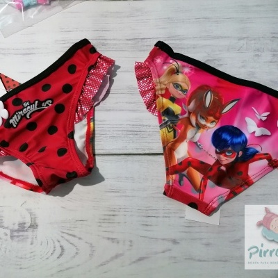 Duas cuecas de banho infantis vermelha e rosa com padrões e personagens, sobre madeira clara