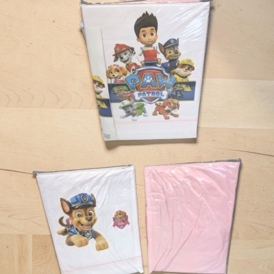Pacotes de roupa de cama infantil com personagens da Patrulha Pata e tecido rosa sobre chão de madeira.