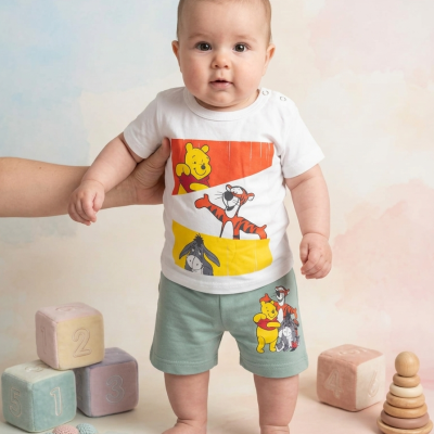 Bebé com conjunto de roupa branca e verde estampada de desenhos animados, rodeado de brinquedos suaves e coloridos.