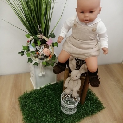 Boneco bebé vestido com jardineira bege sentado numa mala com planta e decoração de coelhos