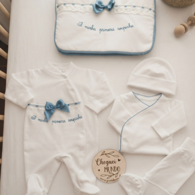 Conjunto de roupa de bebé branca e azul com bordado e laço num fundo claro.