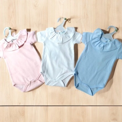 Três bodies para bebé em rosa, branco e azul com gola redonda franzida pendurados em cabides sobre madeira clara.
