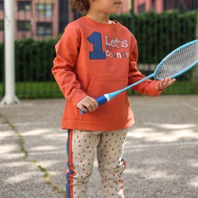 Criança com camisola laranja e calças bege com padrão de letras, a segurar raquete de ténis