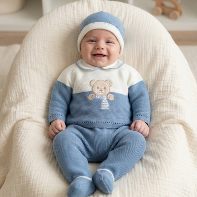 Bebé com conjunto de malha azul e branca com urso bordado sentado em almofada creme.