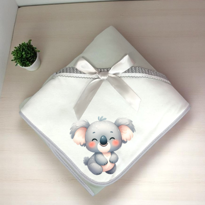 Manta de bebé branca com fita e desenho de coala, sobre mesa de madeira clara com planta em vaso pequeno