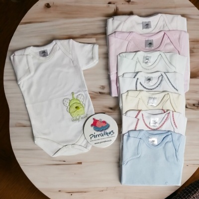 Sete bodies de bebé de várias cores e bordados sobre mesa de madeira com cartão redondo 'Pirralhos'.