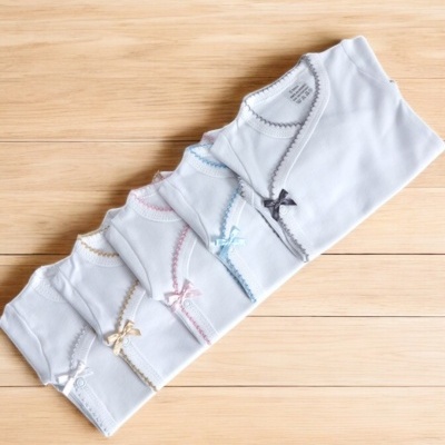 Bodies de bebé brancos com bordas coloridas e laços pequenos sobre superfície de madeira clara.