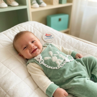 Bebé vestido com jardineira verde de veludo e prendedor de chupeta branco deitado numa cama branca acolchoada