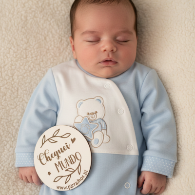 Bebé recém-nascido deitado com babygrow azul e branca e placa redonda de madeira com texto em português.