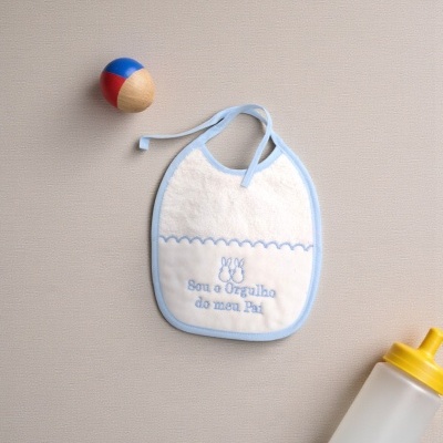Babete branco com bordado azul, brinquedo colorido e biberão com tampa amarela.