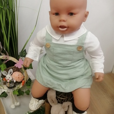 Boneco bebé vestido com camisa branca e vestido verde-água, com botões, meias pretas e sapatinhos brancos, com coelhos de peluche ao lado.