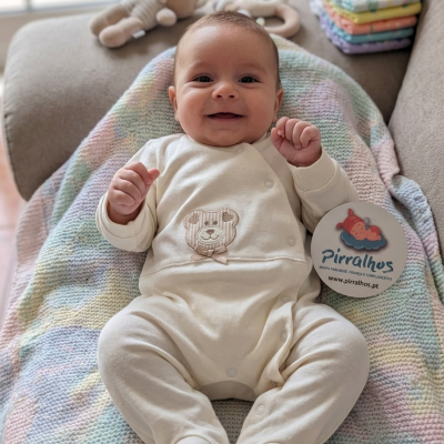 Bebé com macacão creme e manta colorida sobre sofá, com brinquedo e fraldas ao lado