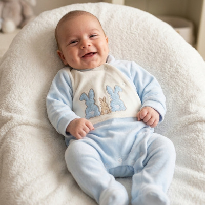 Bebé sorridente sentado em tecido branco macio com fato azul claro e coelhinhos bordados no peito.