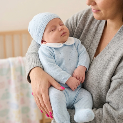 Bebé vestido de azul claro com gorro e macacão, a dormir nos braços de uma mulher com camisola cinzenta, berço ao fundo
