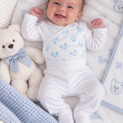 Bebé vestido com roupa branca com padrão azul de Mickey Mouse deitado em mantinha acolchoada azul e branca com urso de peluche ao lado