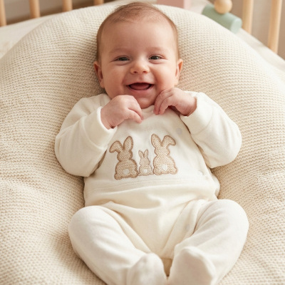 Bebé sorridente vestido com fato creme com coelhos, deitado em almofadão de malha creme num berço de madeira clara.