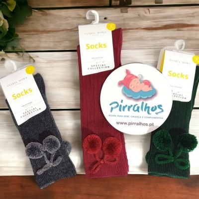 Três pares de meias infantis com pompons em três cores diferentes sobre fundo de madeira clara e flores.