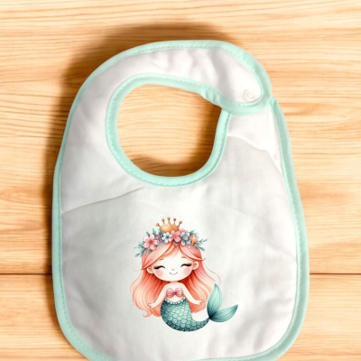 Babete branco com bordas verdes e sereia desenhada, colocado sobre madeira clara.