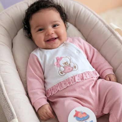 Bebé de roupa rosa e branca com ursinho bordado sentado numa almofada acolchoada bege claro, com etiqueta da marca Pirralhos.