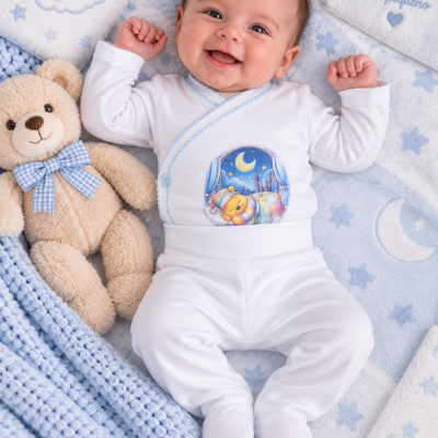 Bebé de pijama branco com estampa colorida de ursinho e lua, deitado em manta azul claro com padrão de estrelas e nuvens ao lado de urso de peluche