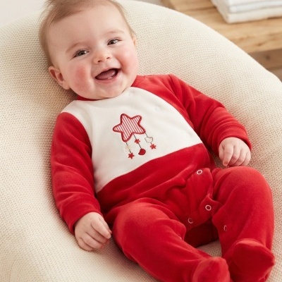 Bebé sorridente vestindo macacão vermelho e branco com estrela decorativa, sentado em almofada creme.