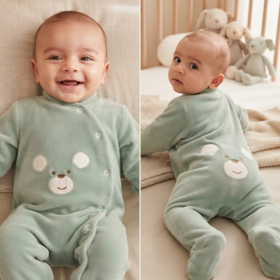 Bebé com pijama verde-água com ursinho bordado numa cama de madeira clara com peluches ao fundo