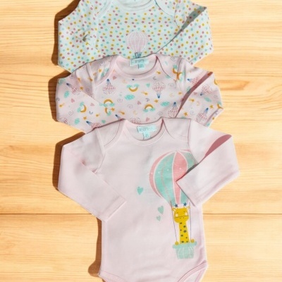 Três bodies bebés coloridos com padrões e ilustrações, mangas compridas, sobre tábua de madeira.