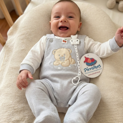 Bebé sorridente em macacão cinzento com ursinho bordado, deitado em almofada creme tricotada, com prende-chupeta e etiqueta Pirralhos.