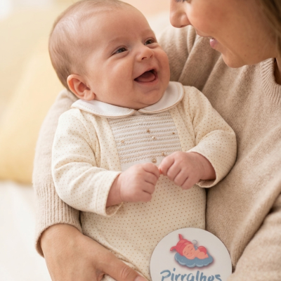 Bebé sorridente com macacão creme e pontos castanhos com etiqueta da marca Pirralhos na mão, segurado por adulto.