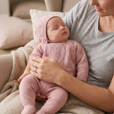 Bebé dormindo com fato de malha rosa muito claro e capuz com botões sentado no colo de mulher