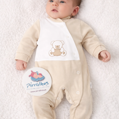 Bebé com macacão bege e branco com ursinho bordado e botões de pressão, sobre fundo branco macio