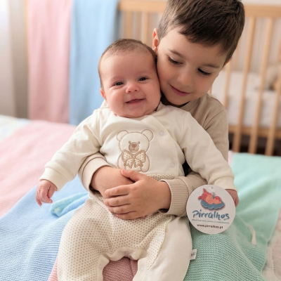 Bebé em macacão creme com ursinho bordado no colo de criança maior, mantas coloridas e berço ao fundo