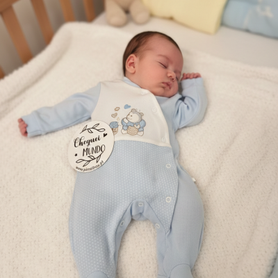 Bebé vestido com macacão azul e branco com desenho de ursinho a dormir numa cama com peluche castanho.