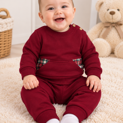 Bebé com fato vermelho bordô e detalhes xadrez sentado em tapete peludo claro com urso de peluche ao fundo.